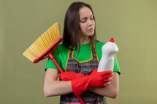 Thinking cleaning young girl wearing uniform in red gloves holding mop looking at cleaning agent on her hand and crossing hands on isolated green wall