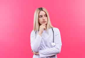 Free photo thinking doctor young blonde girl wearing stethoscope in medical gown put her hand on chin on isolated pink wall