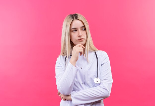 Думающая молодая блондинка со стетоскопом в медицинском халате положила руку на подбородок на изолированной розовой стене