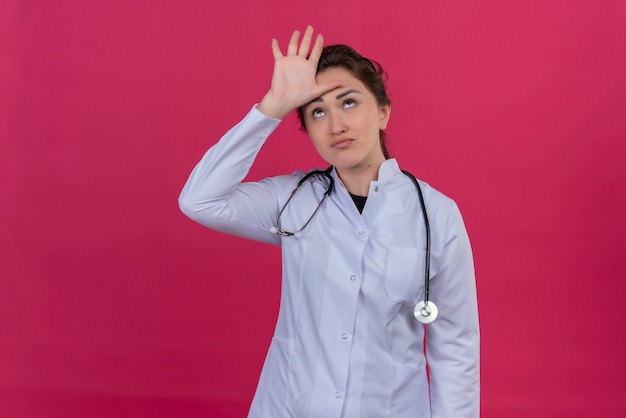 Thinking doctor young girl wearing medical gown and stethoscope put her hand on forehead on isoleted red background