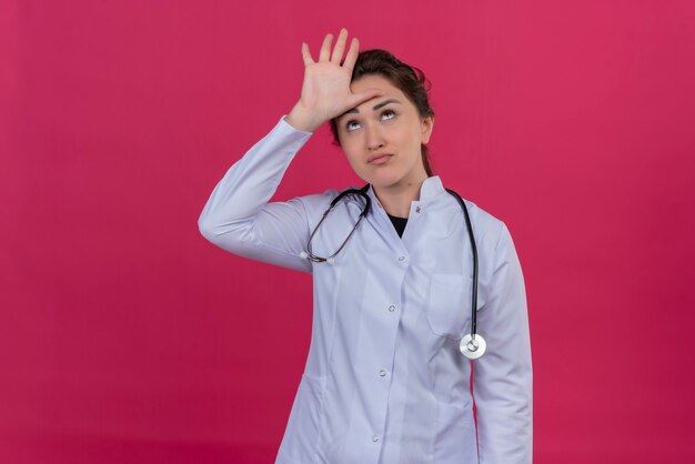 Думающая молодая девушка-врач в медицинском халате и стетоскопе положила руку на лоб на изолированном красном фоне