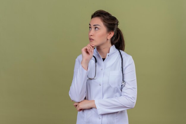 Thinking doctor young girl wearing medical gown wearing stethoscope put her hand under jaw on green background