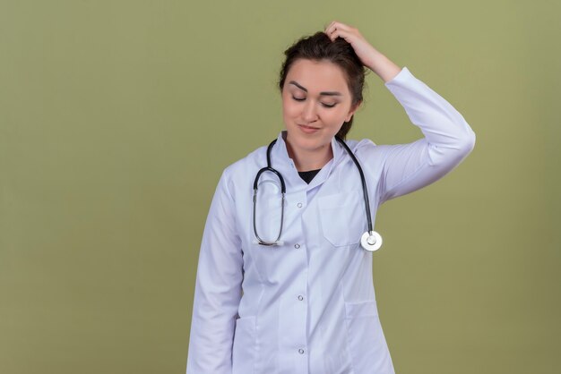 Thinking doctor young girl wearing medical gown wearing stethoscope scratches on green background