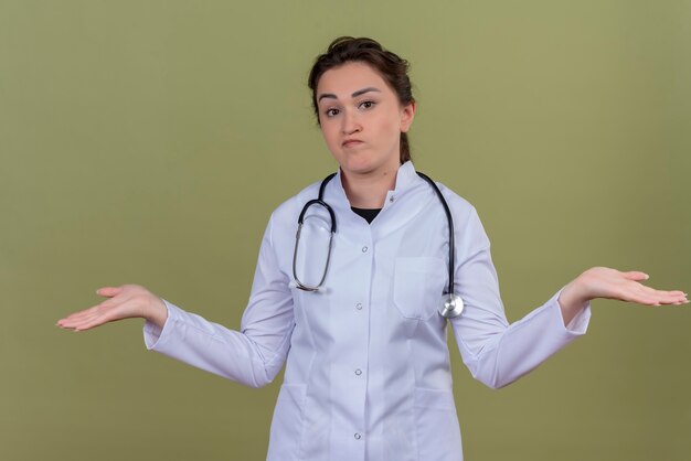 Thinking doctor young girl wearing medical gown wearing stethoscope spreads hands on green background