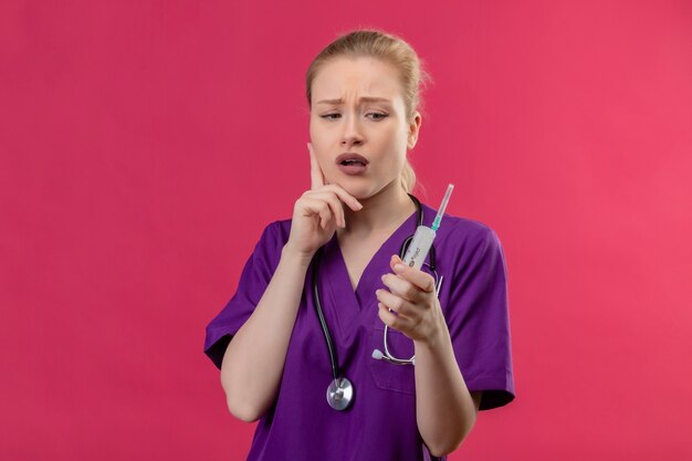 シリンジを保持している聴診器で紫の医療ガウンを着ている医者少女思考思考分離ピンクの背景に頬に手を置く