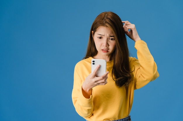 Thinking dreaming young Asia lady using phone with positive expression, dressed in casual clothing feeling happiness and stand isolated on blue background. Happy adorable glad woman rejoices success.