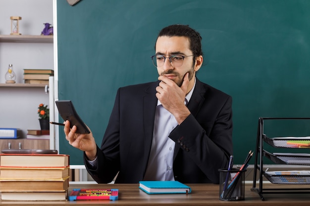 Thinking grabbed chin male teacher wearing glasses holding and looking at calculator sitting at table with school tools in classroom