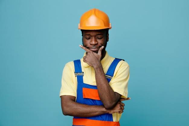 Thinking grabbed chin young african american builder in uniform isolated on blue background