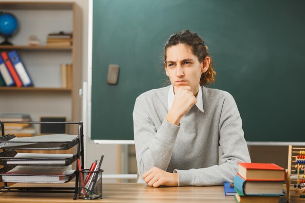 thinking grabbed chin young male teacher sitting at desk with school tools on in classroom