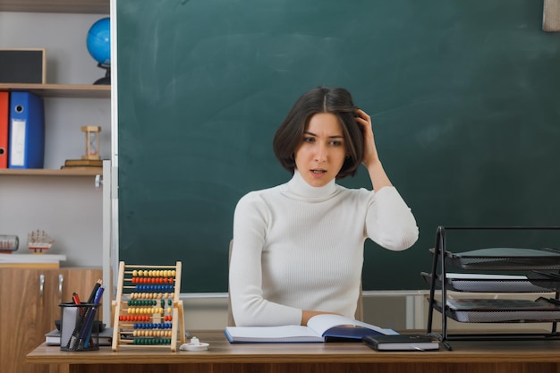 教室で学校の道具をつけて机に座っている頭をつかんだ若い女教師を考えて