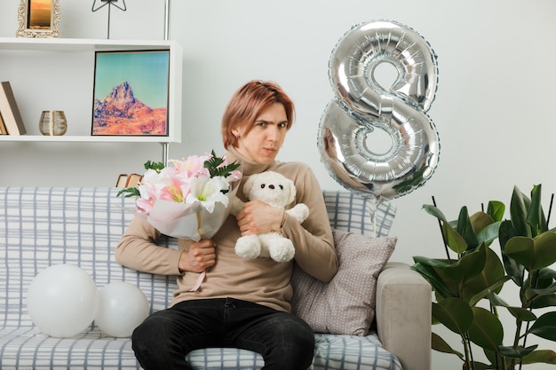 Thinking handsome guy on happy women day holding bouquet with teddy bear sitting on sofa in living room