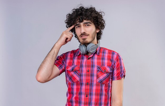 A thinking handsome man with curly hair in checked shirt in headphones pointing at head with index finger 