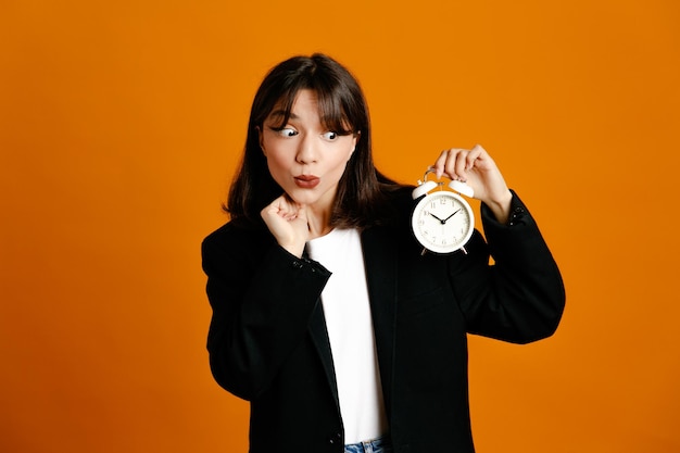 Free photo thinking holding alarm clock young beautiful female wearing black jacket isolated on orange background