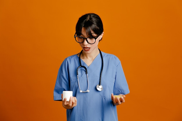 オレンジ色の背景で隔離の均一なフィス聴診器を身に着けているピル若い女性医師の缶を保持することを考えて