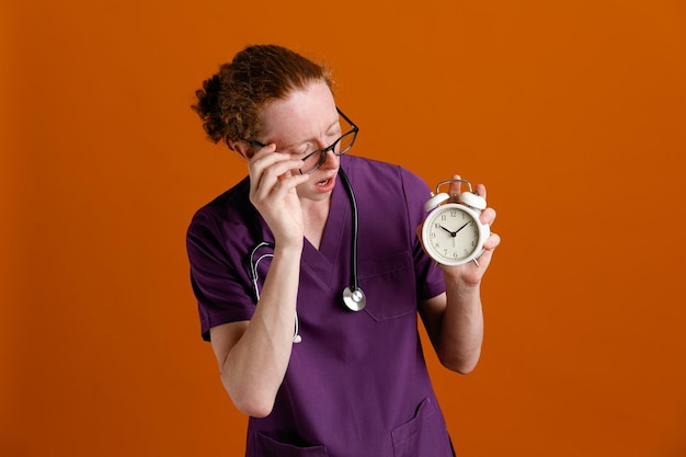 Thinking holding and looking at alarm clock young male doctor wearing uniform with stethoscope isolated on orange background