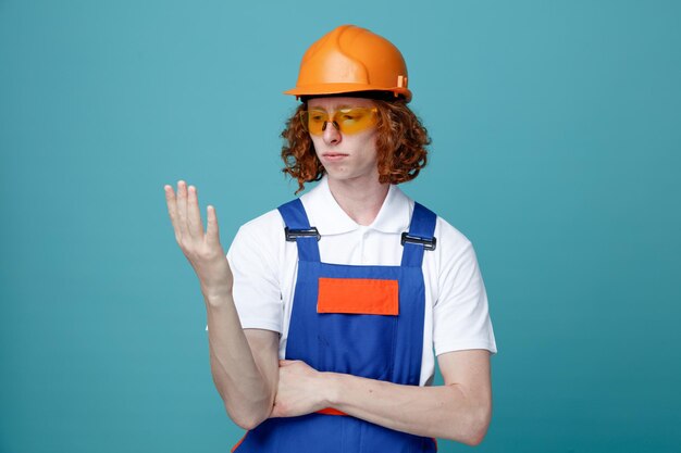 保持し、青の背景に分離された制服を着た若いビルダーの男を手で見て考えてください。