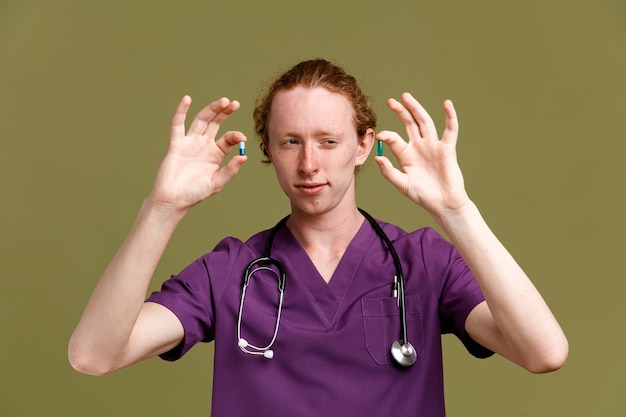думает, держа таблетки, молодой врач-мужчина в униформе со стетоскопом на зеленом фоне