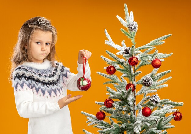 Thinking little girl standing nearby christmas tree wearing tiara with garland on neck holding and points at christmas ball isolated on orange background