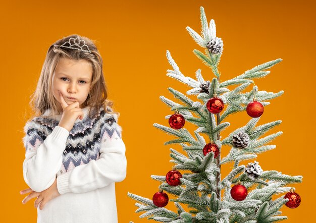 Thinking little girl standing nearby christmas tree wearing tiara with garland on neck putting hand on chin isolated on orange background