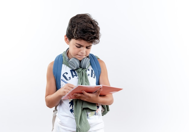 白で隔離のノートに何かを書いているバックバッグとヘッドフォンを身に着けている小さな男子生徒を考えて