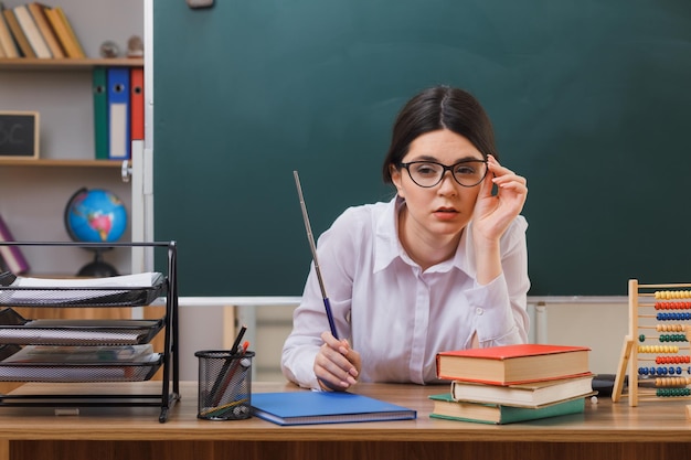 думает, глядя в камеру, молодая учительница в очках, держащая указатель, сидя за партой со школьными инструментами в классе
