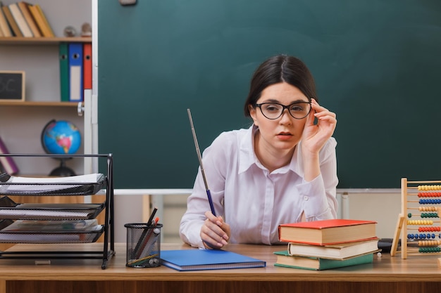 thinking looking at camera young female teacher wearing glasses holding pointer sitting at desk with school tools in classroom