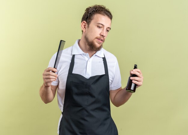 Thinking looking down young male barber wearing uniform holding spray bottle with comb isolated on olive green background