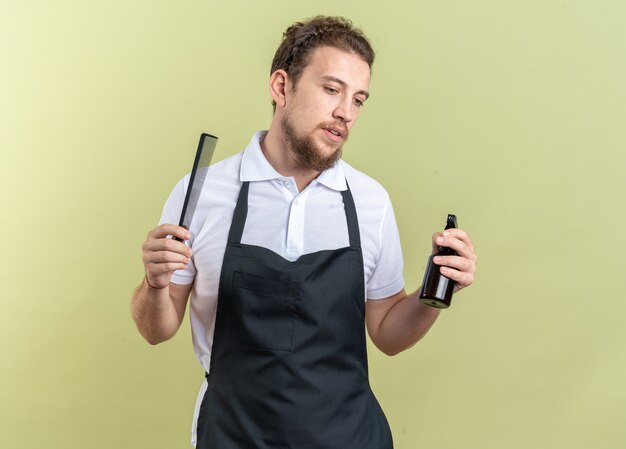 Думая, глядя вниз, молодой мужчина-парикмахер в униформе держит бутылку с распылителем с расческой, изолированной на оливково-зеленом фоне