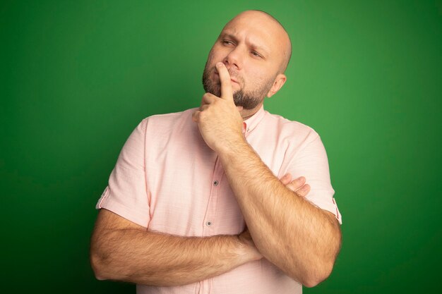 Thinking looking at side middle-aged bald man wearing pink t-shirt putting finger on mouth