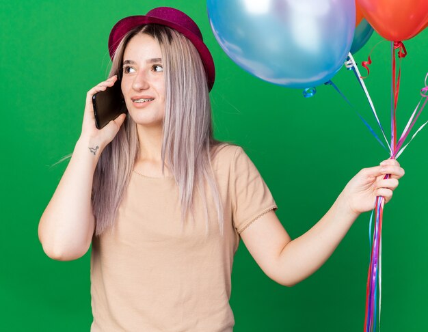 Thinking looking side young beautiful girl wearing party hat and braces holding balloons speaks on phone isolated on green wall