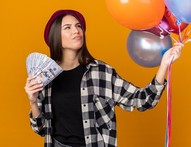Thinking looking side young beautiful girl wearing party hat holding balloons with cash isolated on orange wall
