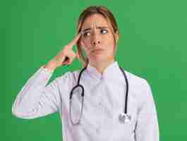 Free photo thinking looking at side young female doctor wearing medical robe with stethoscope putting finger on temple isolated on green wall