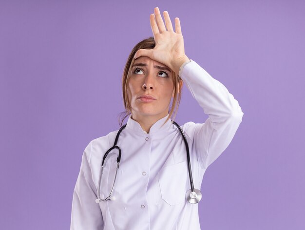 Thinking looking at side young female doctor wearing medical robe with stethoscope showing loser gesture isolated on purple wall