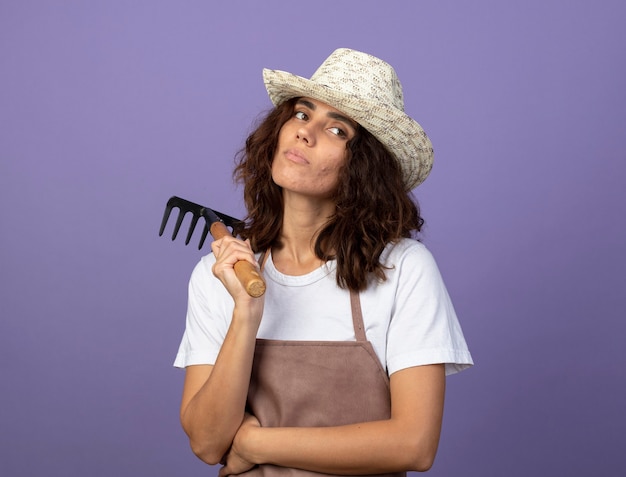 Thinking looking at side young female gardener in uniform wearing gardening hat holding rake on shoulder