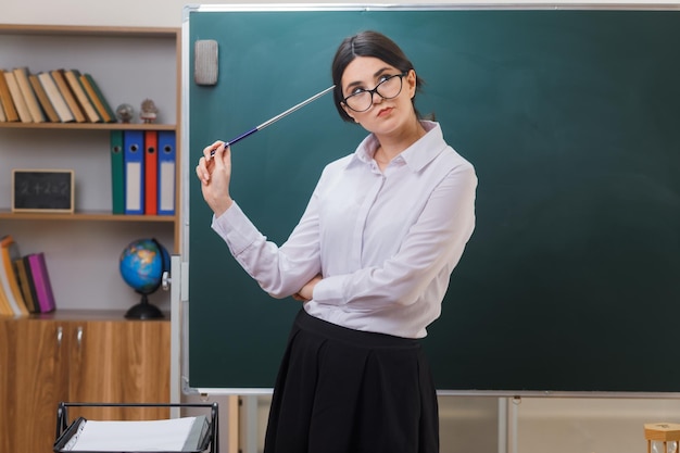 thinking looking at side young female teacher holding pointer on head standing in front blackboard in classroom