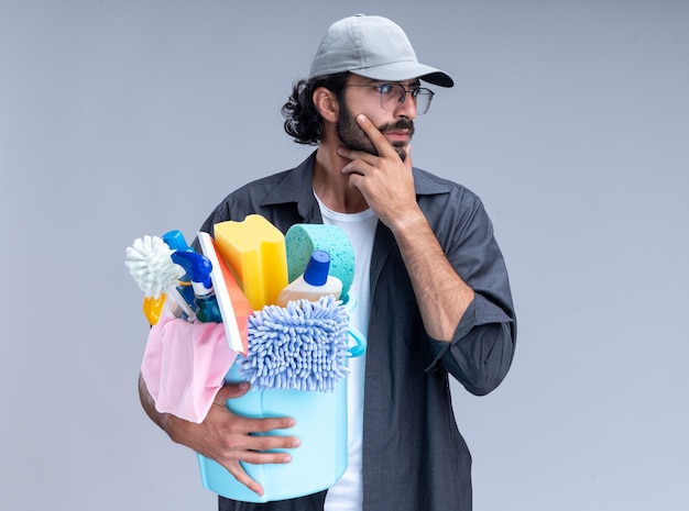 Thinking looking at side young handsome cleaning guy wearing t-shirt and cap holding bucket of cleaning tools grabbed chin isolated on white wall