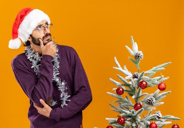 Thinking looking side young handsome guy standing nearby christmas tree wearing christmas hat with garland on neck grabbed chin isolated on orange background