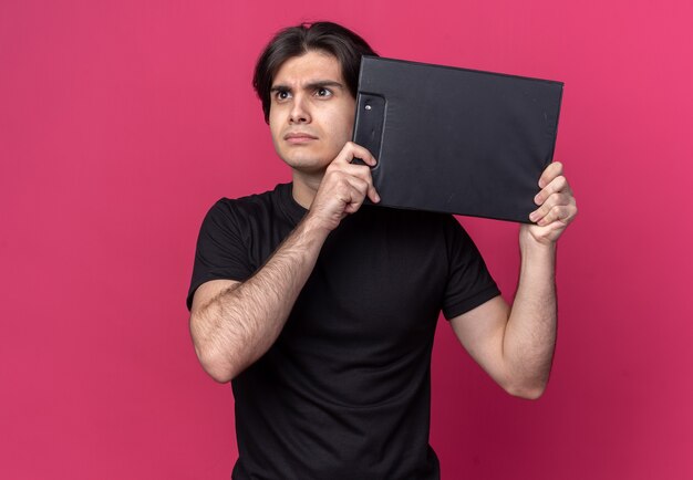 Thinking looking side young handsome guy wearing black t-shirt holding clipboard around face isolated on pink wall