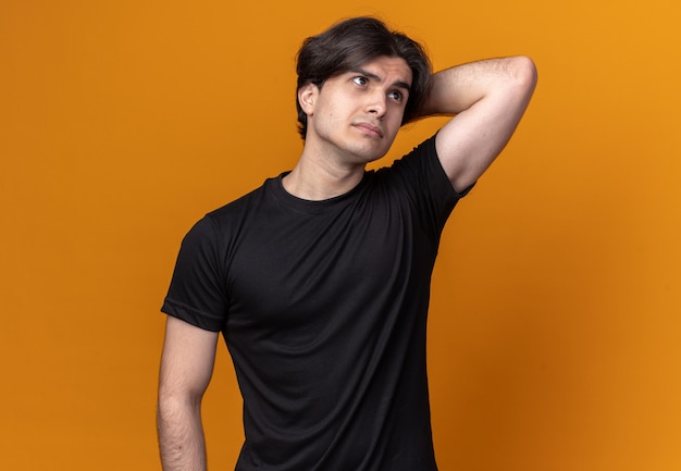 Thinking looking at side young handsome guy wearing black t-shirt putting hand behind head isolated on orange wall