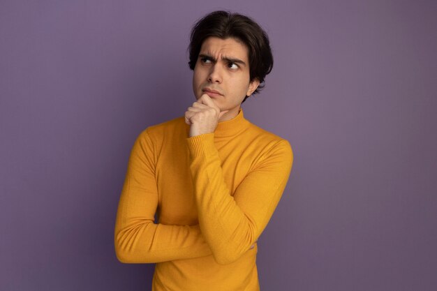 Thinking looking at side young handsome guy wearing yellow turtleneck sweater grabbed chin isolated on purple wall with copy space