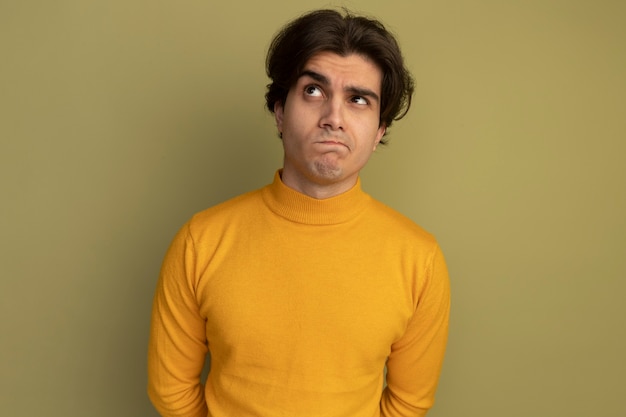 올리브 녹색 벽에 고립 된 노란색 터틀넥 스웨터를 입고 측면 젊은 잘 생긴 남자를보고 생각