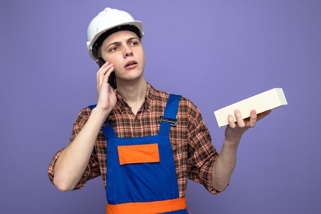 Думая, смотрящая сторона, молодой мужчина-строитель в униформе, держащий кирпич, разговаривает по телефону
