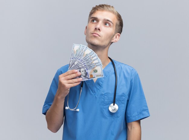 Думая, глядя на сторону молодого мужчины-доктора в униформе врача со стетоскопом, держащего деньги, изолированные на белой стене
