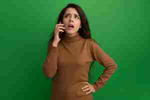 Free photo thinking looking up young beautiful girl speaks on phone putting hand on hip isolated on green wall