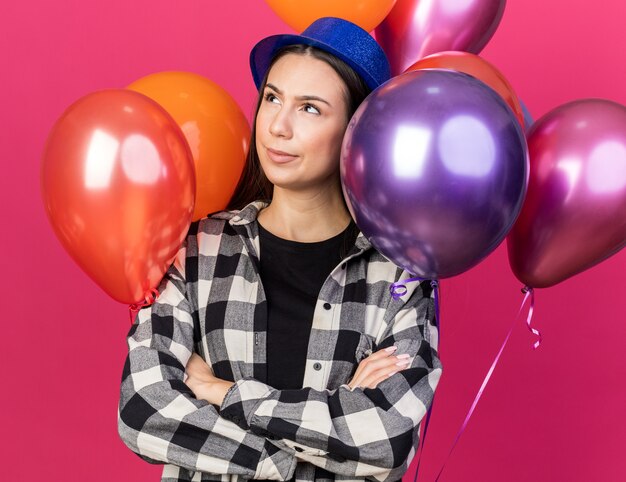 Thinking looking up young beautiful woman wearing party hat standing in front balloons crossing hands isolated on pink wall