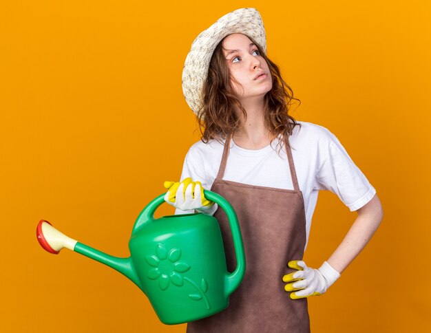 Thinking looking up young female gardener wearing gardening hat and gloves holding watering can putting hand on hip isolated on orange wall