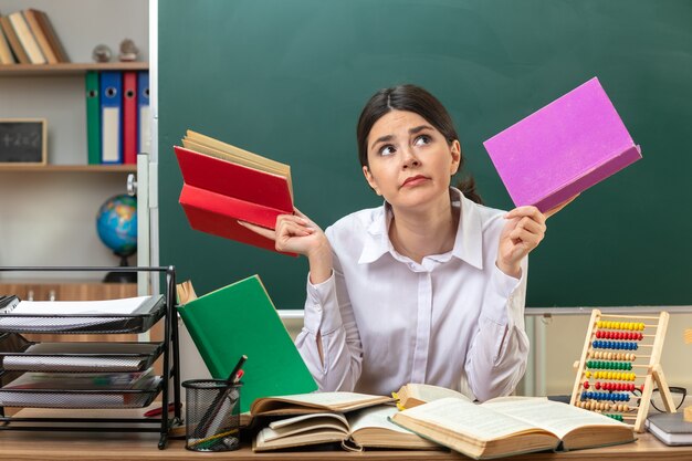 Thinking looking up young female teacher holding book sitting at table with school tools in classroom