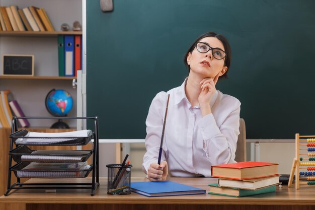教室で学校の道具を持って机に座っているポインターを保持している眼鏡をかけている若い女教師を見上げることを考えて
