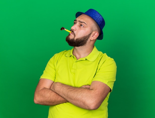 Thinking looking up young man wearing blue party hat blowing party whistle crossing hands isolated on green wall