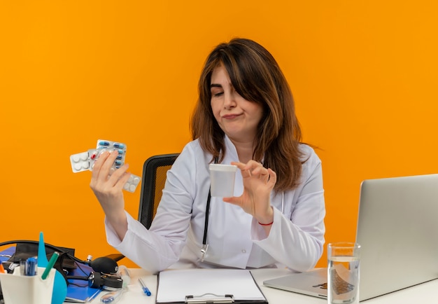 Free photo thinking middle-aged female doctor wearing medical robe with stethoscope sitting at desk work on laptop with medical tools holding and looking at empty can with pills on isolated orange wall